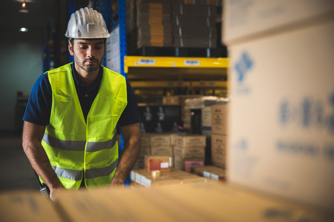 Warehouse employee moving boxes in a vest and a helmet but in need of impact resistant gloves as part of his ppe.