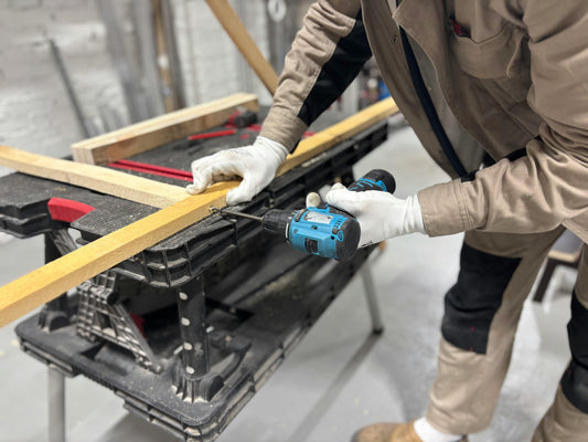 A person wearing work gloves and a beige utility uniform uses a cordless power drill to drive a screw into a wooden board on a black workbench, with additional lumber pieces and tools visible in the background of the workshop.