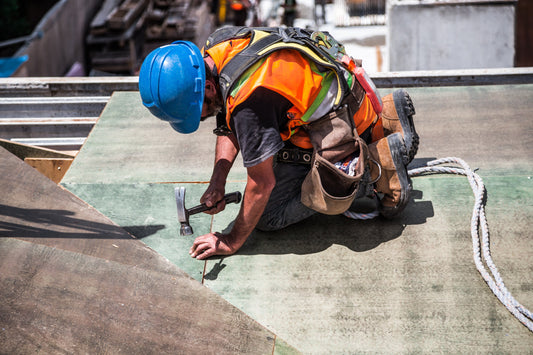 Construction worker in vest and hard hat hammering a nail on the roof of a building with no work gloves on his hands