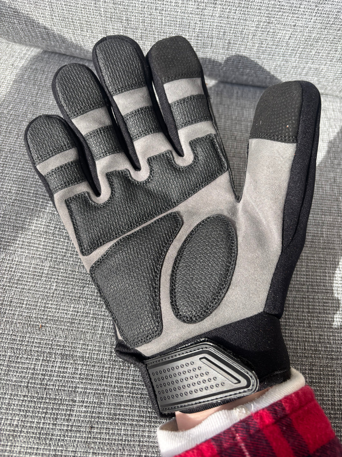 A single work glove shown flat against a gray textured fabric background. The glove is primarily light gray with black textured panels on the fingers, palm, and wrist strap, plus a small red-and-white plaid cuff visible at the bottom edge. Safety Tuff gloves with finger guards (finger caps).